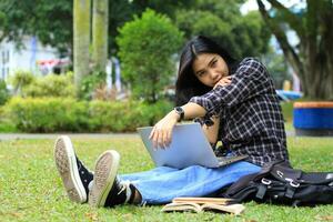 Lycklig asiatisk studerande läsning en bok och använder sig av bärbar dator till studie för befordran, se till kamera foto