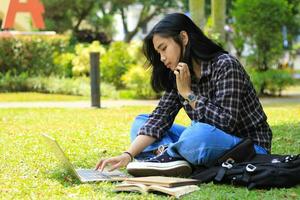 porträtt av fokuserade ung asiatisk kvinna surfing internet använder sig av bärbar dator till arbetssätt avlägset, läsa bok, och skrivning i anteckningsbok. attraktiv asiatisk studerande studie i utomhus foto