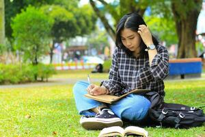 skön asiatisk ung kvinna högskola studerande fokuserade skrivning på anteckningsbok och läsning bok i utomhus stad parkera foto