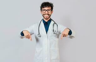 ung läkare pekande en befordran ner. stilig läkare pekande ner ett isolerat reklam Plats. Lycklig läkare pekande fingrar ner foto