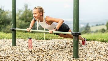 sportig ung kvinna arbetssätt ut gymnastik i ett utomhus- Gym använder sig av parallell barer. skön blond flicka med flätor Arbetar ut utomhus håller på med armhävningar på ett upphöjd bar foto