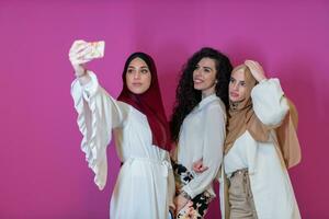 grupp av skön muslim kvinnor två av dem i modern klänning med hijab använder sig av mobil telefon medan tar selfie bild isolerat på rosa bakgrund representerar modern islam mode teknologi foto
