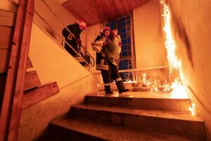 modig brandman sänker sig trappa av en brinnande byggnad och innehar sparade flicka i hans vapen. öppen brand och ett brandman i de bakgrund. foto