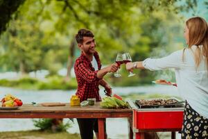 Lycklig par toasting röd vin glas medan har picknick franska middag fest utomhus- under sommar Semester semester nära de flod på skön natur foto