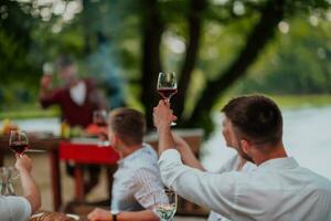 grupp av Lycklig vänner toasting röd vin glas medan har picknick franska middag fest utomhus- under sommar Semester semester nära de flod på skön natur foto