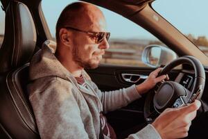 en man med en solglasögon körning en bil och typ en meddelande på smartphone på solnedgång. de begrepp av bil resa foto