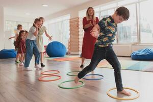 små barnkammare skola barn med kvinna lärare på golv inomhus i klassrum, håller på med träning. Hoppar över hula ring cirklar Spår på de golv. foto