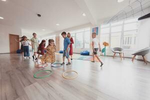 små barnkammare skola barn med kvinna lärare på golv inomhus i klassrum, håller på med träning. Hoppar över hula ring cirklar Spår på de golv. foto