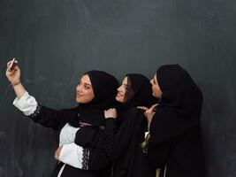 grupp av ung skön muslim kvinnor i modern klänning med hijab använder sig av smartphone medan tar selfie bild i främre av svart bakgrund foto