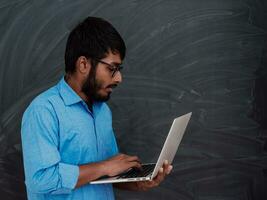 indisk leende ung studerande i blå skjorta och glasögon använder sig av bärbar dator och Framställ på skola svarta tavlan bakgrund foto