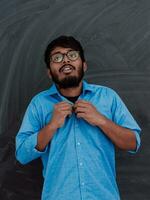 indisk ung studerande bär glasögon sätter på en blå skjorta medan stående i främre av de skola svarta tavlan foto