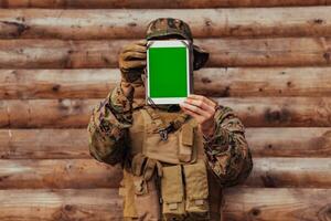 soldat använder sig av läsplatta dator mot gammal trä- vägg i militär läger till stanna kvar i Kontakt med vänner och familj foto