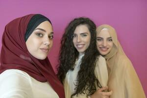 grupp av skön muslim kvinnor två av dem i modern klänning med hijab använder sig av mobil telefon medan tar selfie bild isolerat på rosa bakgrund representerar modern islam mode teknologi foto