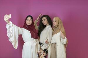 grupp av skön muslim kvinnor två av dem i modern klänning med hijab använder sig av mobil telefon medan tar selfie bild isolerat på rosa bakgrund representerar modern islam mode teknologi foto
