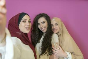 grupp av skön muslim kvinnor två av dem i modern klänning med hijab använder sig av mobil telefon medan tar selfie bild isolerat på rosa bakgrund representerar modern islam mode teknologi foto