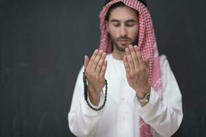 en ung arab man i traditionell kläder framställning en traditionell bön till Gud håller hans händer i bön- gest i främre av en svart bakgrund foto