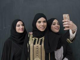 grupp av ung skön muslim kvinnor i modern klänning med hijab använder sig av smartphone medan tar selfie bild i främre av svart bakgrund foto