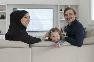 Lycklig muslim familj med dotter kvinna i traditionell modern klänning har roligt och Bra tid tillsammans medan Sammanträde på soffa foto