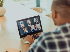 afrikansk amerikan man i glasögon Sammanträde på en tabell i en modern levande rum, använder sig av en bärbar dator för företag video chatt, konversation med vänner och underhållning foto