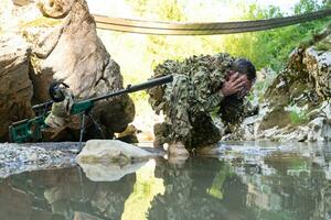 soldat i en kamouflage kostym enhetlig dricka färsk vatten från de flod. militär prickskytt gevär på de sida. foto