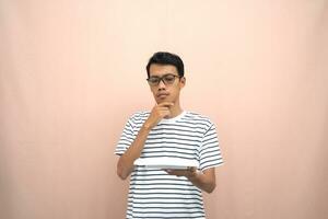 porträtt av ett asiatisk man i glasögon bär en tillfällig randig t-shirt. bärande ett tömma tallrik medan tänkande. beige bakgrund. foto