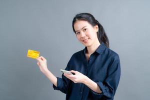 porträtt av en glad ung asiatisk tjej som visar plastkreditkort medan du håller mobiltelefonen på grå bakgrund