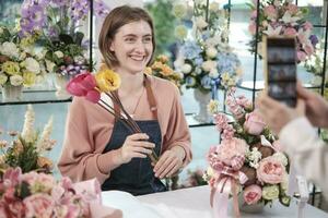 e-handel företag, ett ung vit kvinna blomsterhandlare demonstrerar och visar blommig arrangemang via uppkopplad leva strömning med smartphone Ansökan i ljus blomma affär, skön blommar Lagra. foto