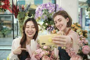 e-handel företag partner, två ung kvinna blomsterhandlare arbetare visa blommig arrangemang, uppkopplad leva strömning, och selfies med smartphone Ansökan i ljus blomma affär, skön blommar Lagra. foto