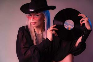 sexig dj kvinna utseende över färgad glasögon innehav en vinyl spela in med en svart hatt. foto