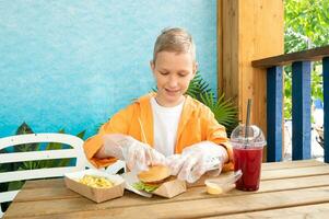 en söt pojke i en gata Kafé äter snabb mat med citronsaft i plast disponibel handskar foto