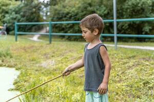 liten leende barn spelar med en pinne förbi en damm i en offentlig parkera foto