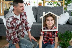 Ofullständig familj pappa och dotter är njuter ny Hem, Sammanträde på soffa. inteckning, försäkring och skydd, uppköp och rör på sig in i din egen Hem, grön hus med inlagd växt foto