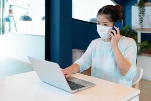 kvinna bär mask och använder dator för videosamtal på arbetsplats.