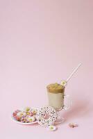 dalgona kaffe i en transparent kopp med en papper rör, en tallrik med blommor daisy och en utsökt aptitretande donera med rosa glasyr. kopia Plats foto