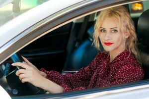 skön framgångsrik blond kvinna i en röd klänning Sammanträde i en bil Bakom de hjul foto