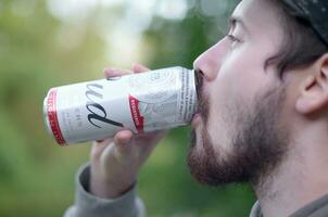 ung skäggig man drycker budweiser knopp öl i aluminium kan på grön träd suddig bakgrund foto