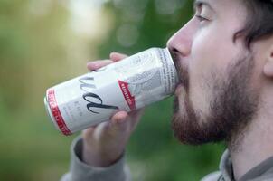 ung skäggig man drycker budweiser knopp öl i aluminium kan på grön träd suddig bakgrund foto