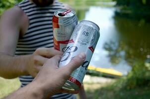 ung man höja budweiser knopp öl kan med manlig vän på suddig flod med kajak och träd foto