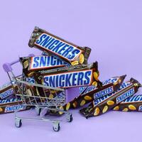 många snickers choklad barer staplade stänga upp med handla vagn på ljus violett bakgrund. snickers barer är produceras förbi fördärvar inkorporerad foto