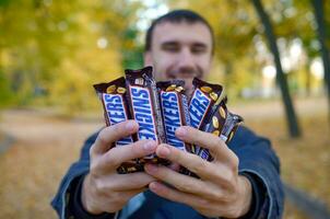 Kharkov, ukraina - oktober 8, 2019 en ung caucasian skäggig man visar många snickers choklad barer i brun omslag i höst parkera. snickers choklad tillverkad förbi fördärvar foto