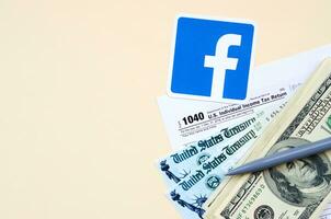Facebook tryckt logotyp lögner med 1040 enskild inkomst beskatta lämna tillbaka form med återbetalning kolla upp och hundra dollar räkningar på beige bakgrund. hjälp i beskatta period från social nätverk begrepp foto