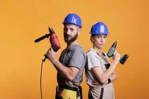 självsäker renovatörer stående med elektrisk kraft borrar, ser på kamera och verkande självsäker i studio. team av byggare innehav borrning pistol verktyg till arbete på renovering, renovering. foto