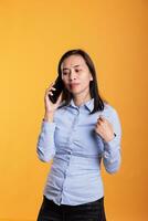glad asiatisk kvinna talande på telefon linje, svar telefon ring upp över gul bakgrund. modell använder sig av smartphone till ha avlägsen konversation med anställd, chattar på mobil telefon i studio. foto