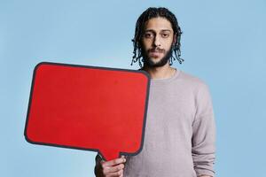 arab man innehav röd tömma baner med plats för text för reklam falsk upp och ser på kamera. modell stående med tom dialog ram för meddelande med kopia Plats porträtt foto