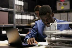 affärsman analyserar administrativ filer, sökande för byråkrati spela in i företags- depositarie. afrikansk amerikan bokhållare arbetssätt sent på natt på bokföring Rapportera foto
