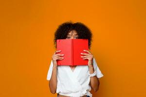 fundersam afrikansk studerande kvinna med bok peta inomhus- över orange vägg. foto