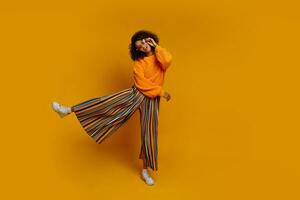 söt afrikansk flicka i orange Tröja och färgrik byxor Hoppar och rolig dans på gul bakgrund. full längd. foto