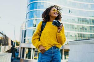 framgångsrik svart kvinna fotograf framställning foton på modern arkitektur bakgrund. bär tillbaka packa, gul Tröja och glasögon.