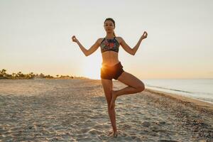 en kvinna i en sporter behå och shorts är håller på med yoga på de strand foto