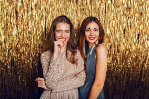 stänga upp studio bild av två Fantastisk sexig fira flickor med röd mun, överraskning ansikte, skrattande , Framställ på gyllene glittrande bakgrund. ny år fest humör. foto